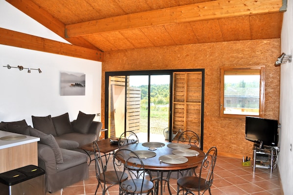 Une pièce chaleureuse avec un plafond en bois, un canapé, une table à manger avec des chaises, un téléviseur et une vue sur l’extérieur grâce à une grande fenêtre.