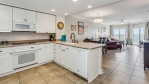 Una cocina moderna con armarios blancos, un microondas integrado y una isla grande. Hay un área de comedor con un sofá y una mesa, y un ventanal con vistas.