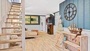 Un salon moderne avec un escalier en bois, une grande horloge murale et une étagère décorative ornée de figurines de bateaux.