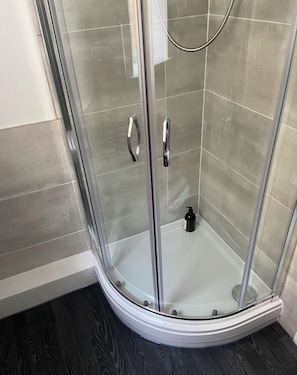 A glass-enclosed shower with a curved floor, a handheld showerhead, and a black soap dispenser on the floor.