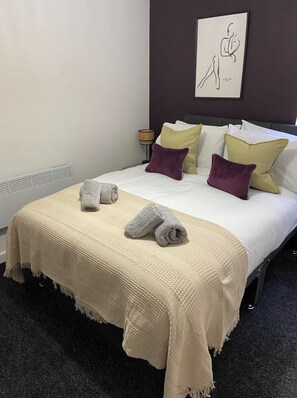 A neatly made bed with a beige blanket and two rolled towels, a bedside lamp, and a wall art piece.