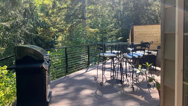 Una terraza de madera con una parrilla, mobiliario exterior y vistas a árboles frondosos.