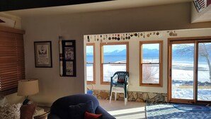 A living room with a blue armchair, a lamp, and a stone wall. There are framed pictures on the wall and a window with a view of mountains and a snowy landscape. - Gorgeous, sweeping views of Lake Tahoe from 3 story lake front condo! (Tahoe City)