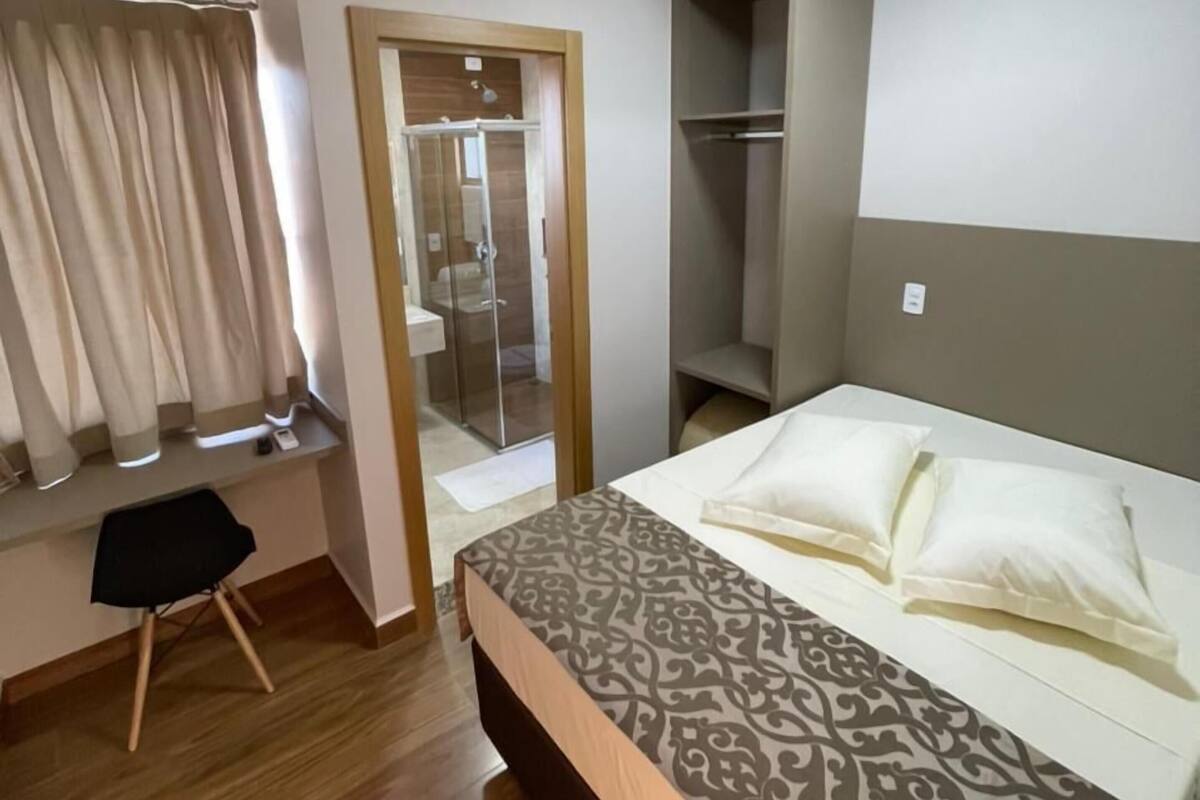 Habitación de hotel con cama, escritorio, silla, armario y baño visible a través de una puerta de vidrio.