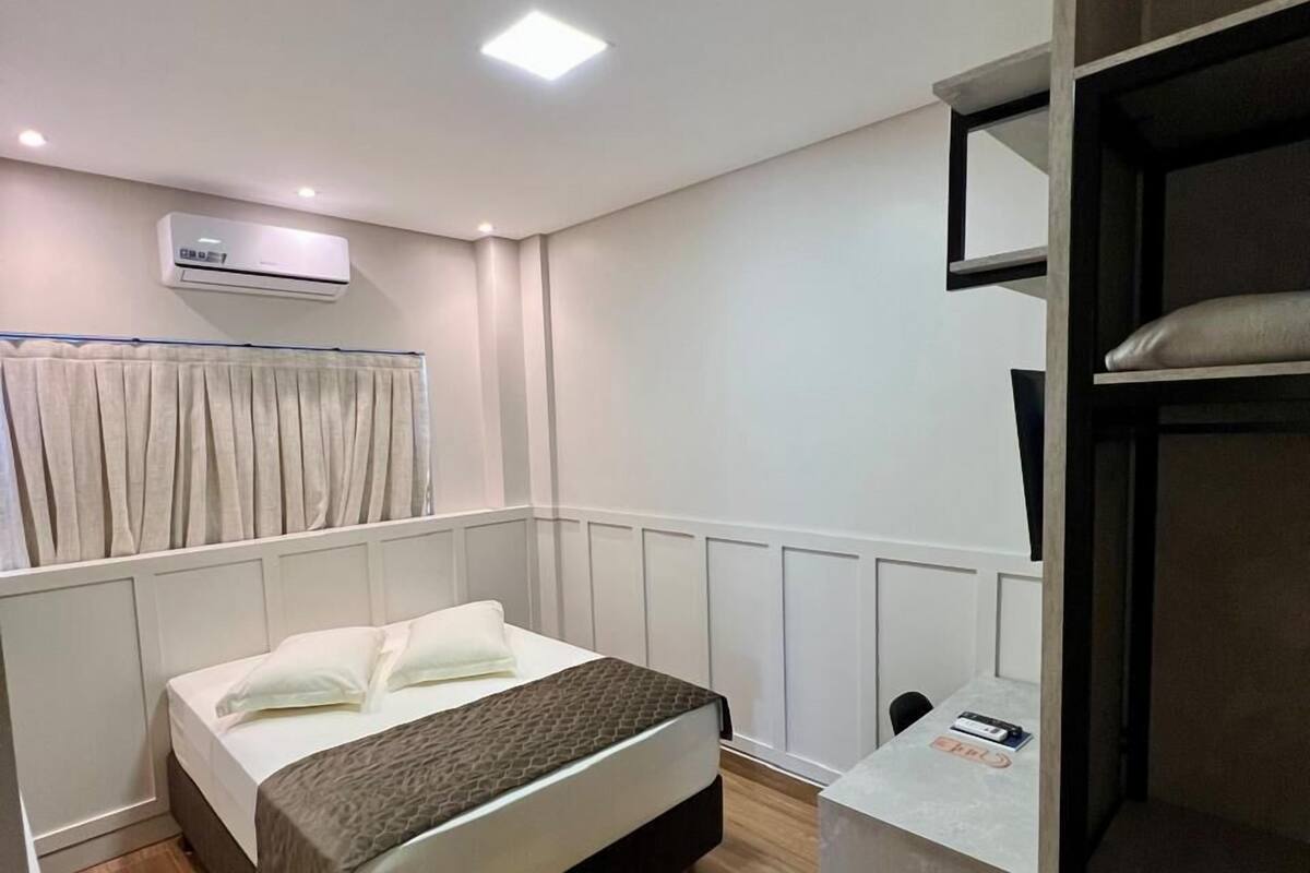 Un dormitorio con cama, escritorio, silla, unidad de aire acondicionado y ventana con cortinas.