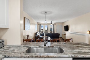 A kitchen with a marble countertop, a stainless steel sink, wooden chairs, and a dining table.