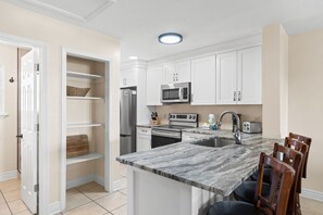 A modern kitchen with white cabinets, a stainless steel refrigerator, a microwave, an oven, and a sink. There is a marble countertop and wooden chairs.