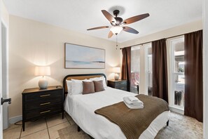 A hotel bedroom with a bed, bedside tables, a ceiling fan, a window with curtains, and a painting on the wall.