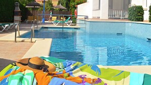 A swimming pool with lounge chairs and umbrellas, a tiled deck, and a person relaxing on a colorful towel.