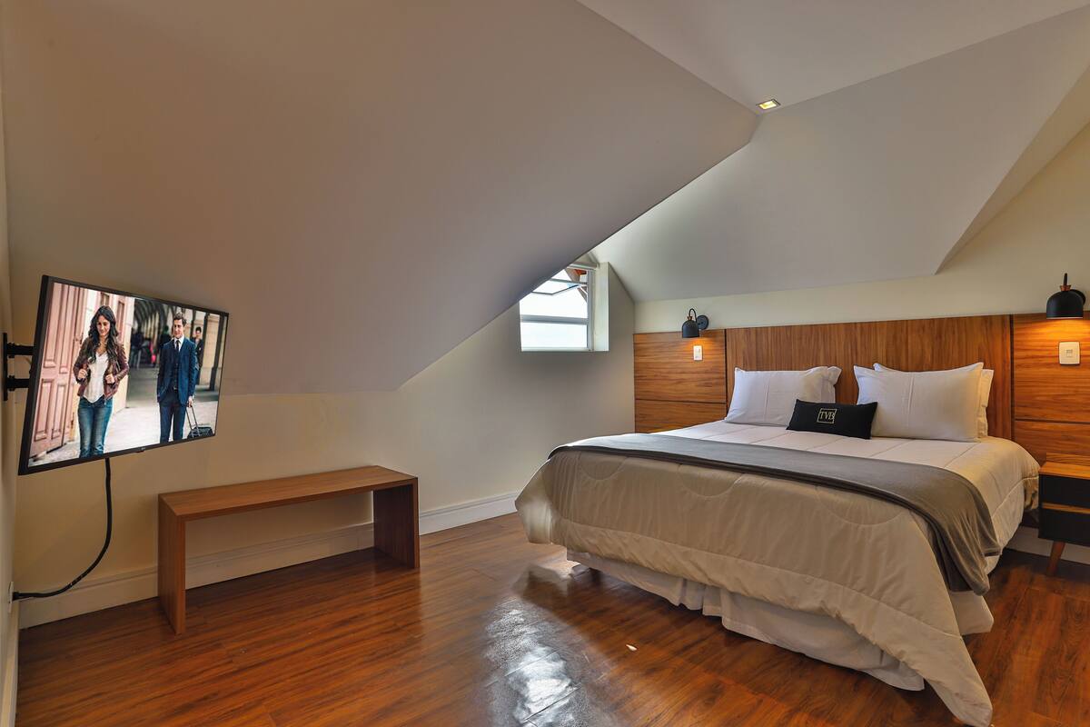 Un dormitorio con techo inclinado, una cama grande con ropa blanca, cabecera de madera, un banco y un televisor montado en la pared.