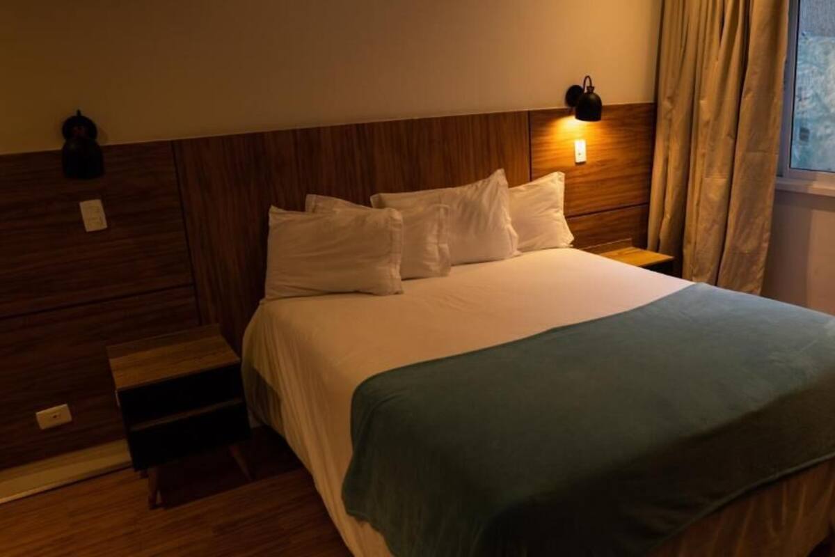 Una cama bien hecha con sábanas blancas y una manta verde, cabecera de madera, luz de noche en la pared y una ventana con cortinas.