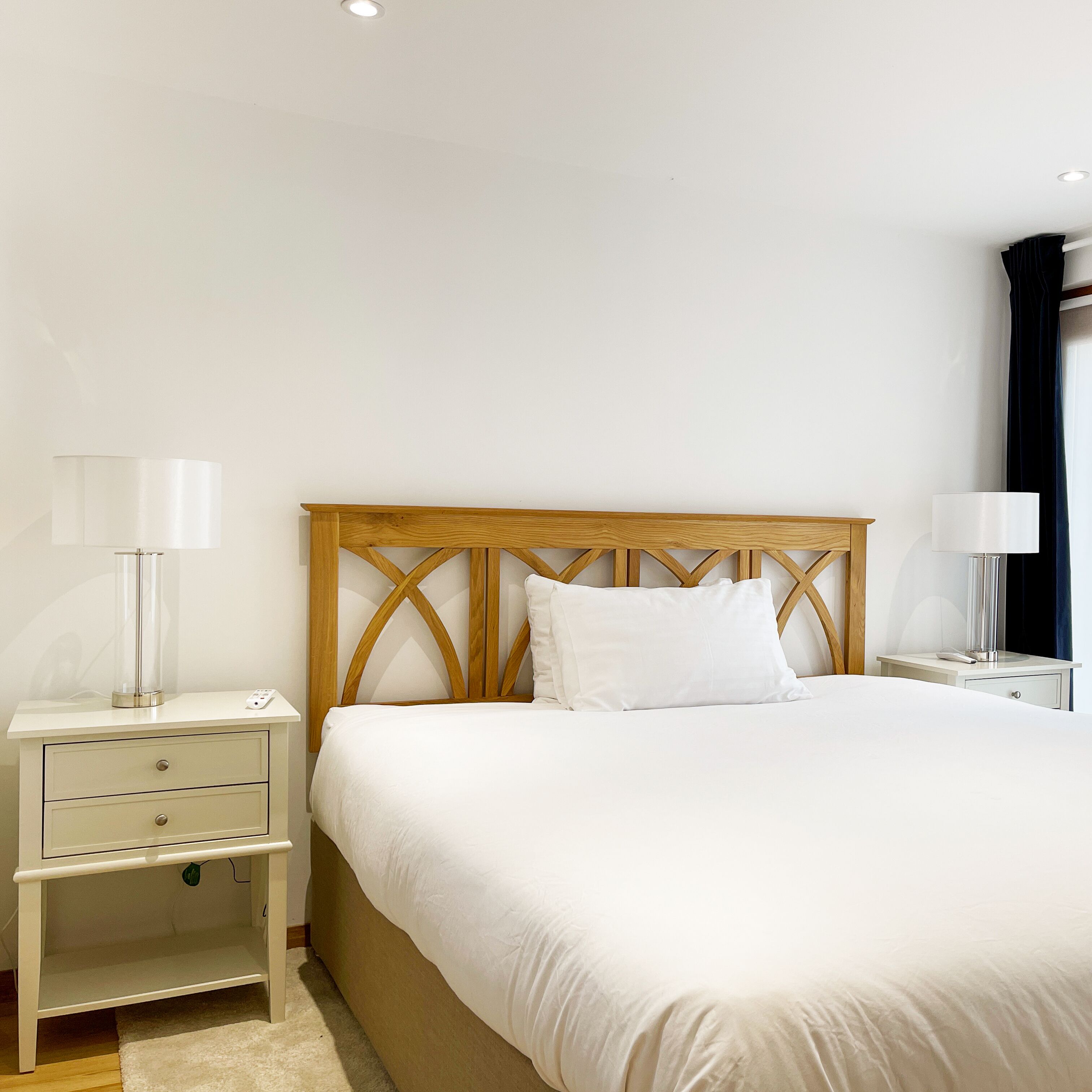 A neatly arranged hotel room with a wooden headboard, a large bed, two bedside tables with lamps, and a small drawer unit.