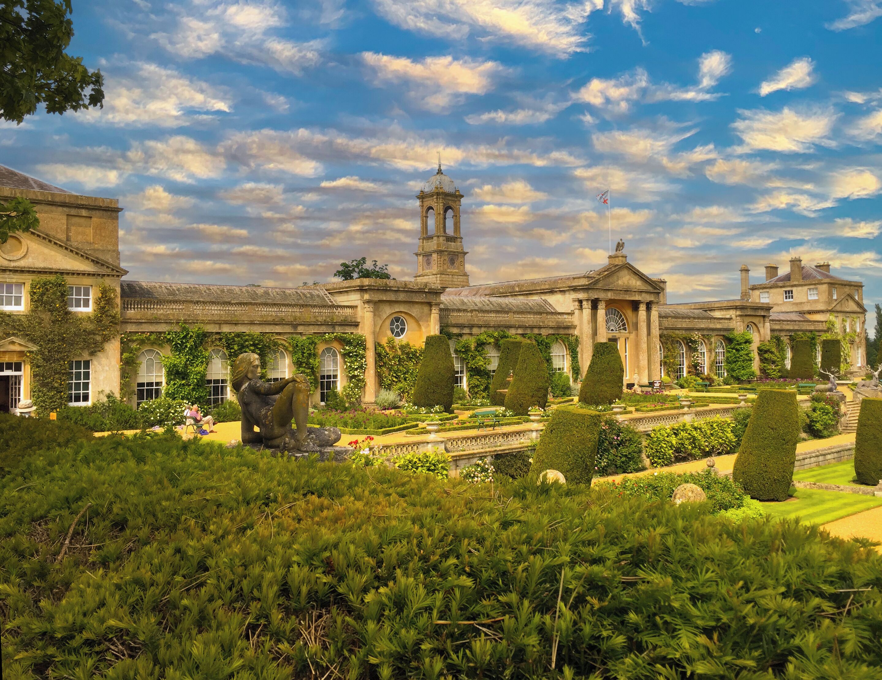 A historic building with a central tower, arched windows, and a formal garden with trimmed hedges and statues.