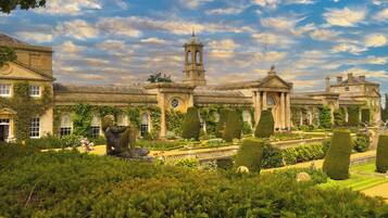 A historic building with a central tower, arched windows, and a formal garden with trimmed hedges and statues.