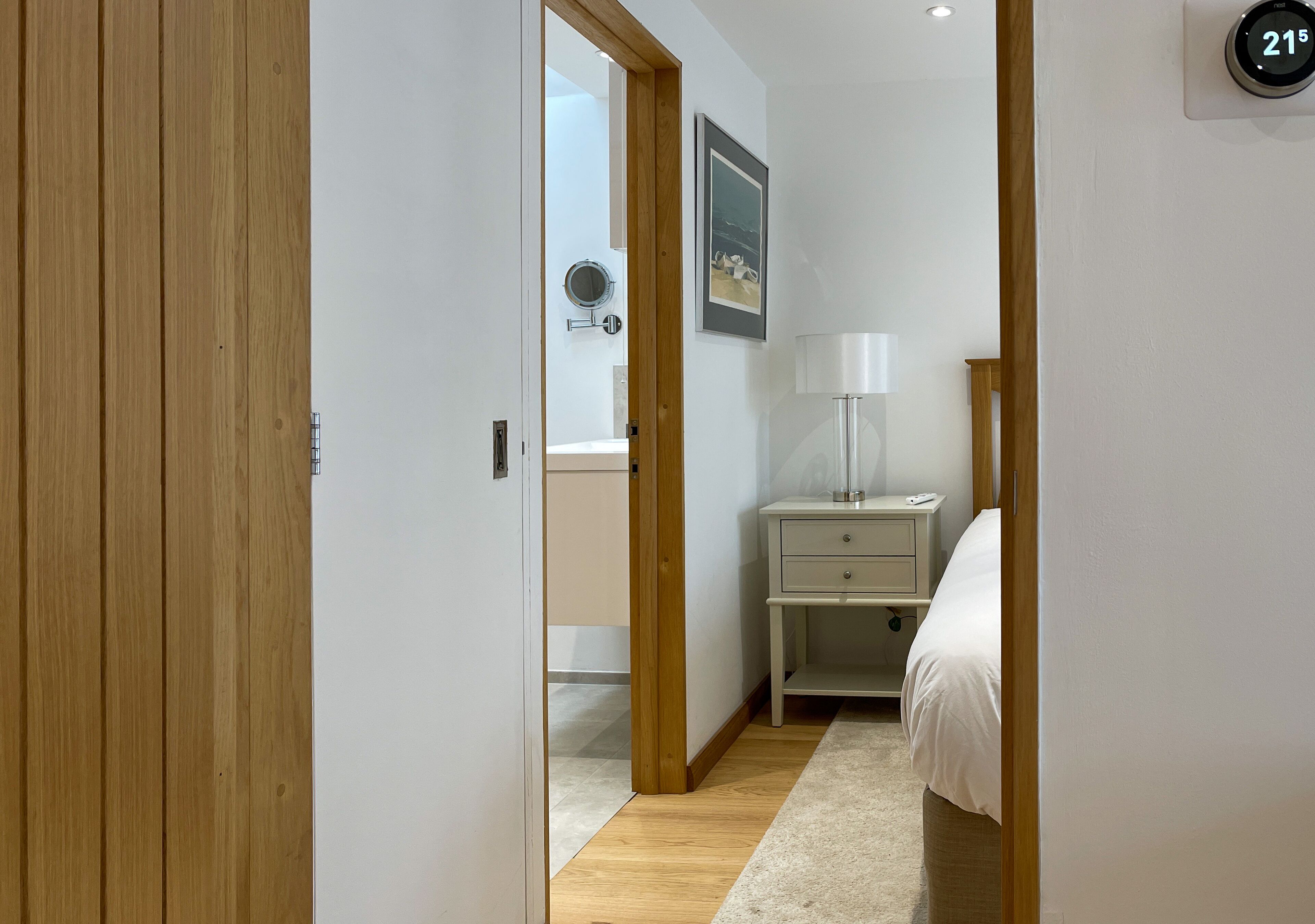 A hotel room with a wooden door, a bedside table with a lamp, a bed, and a wall-mounted thermostat.