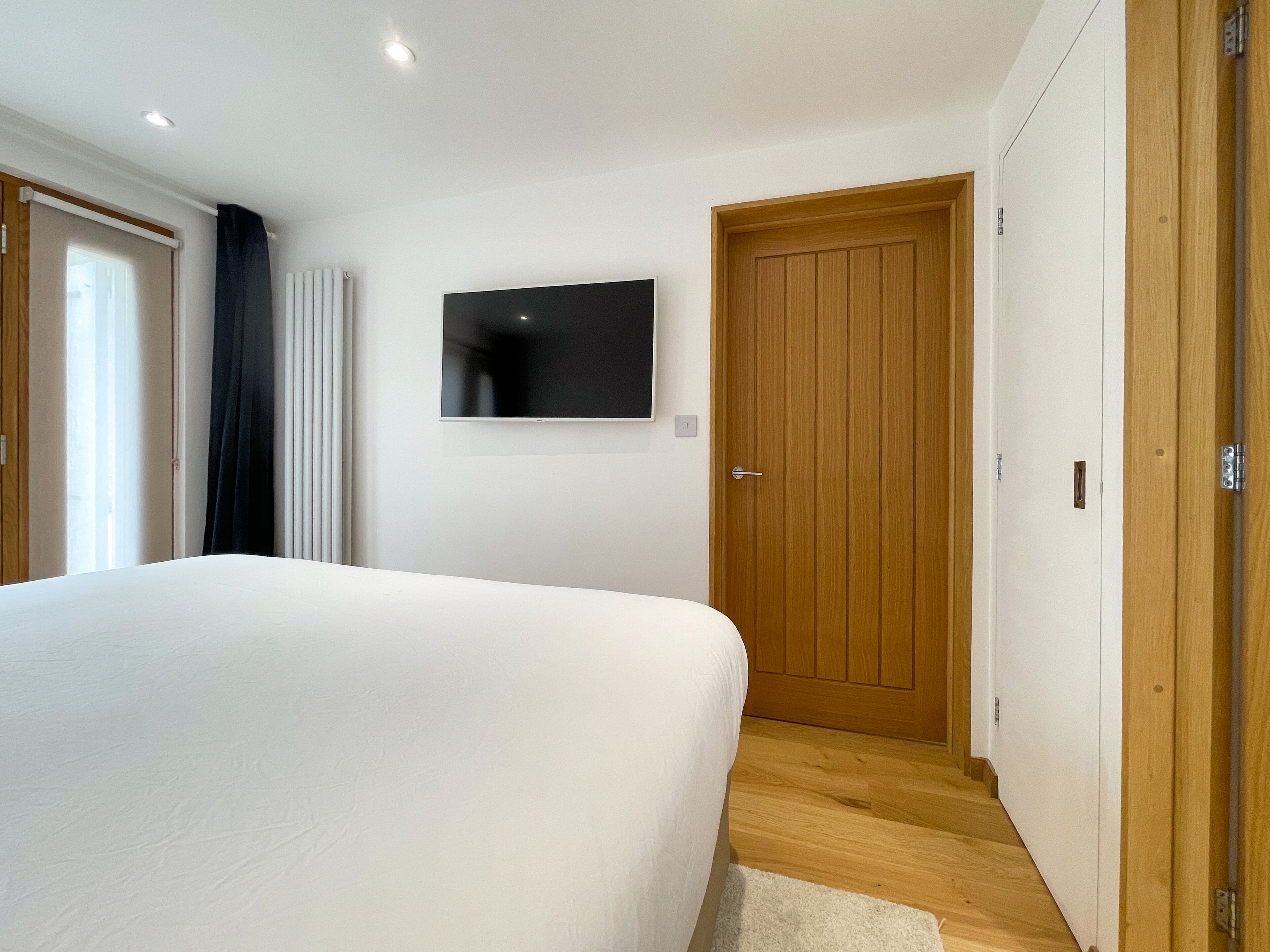 Ein Schlafzimmer mit einem großen Bett, einem Holzschrank, einem an der Wand befestigten Fernseher und einem Fenster mit Vorhängen.