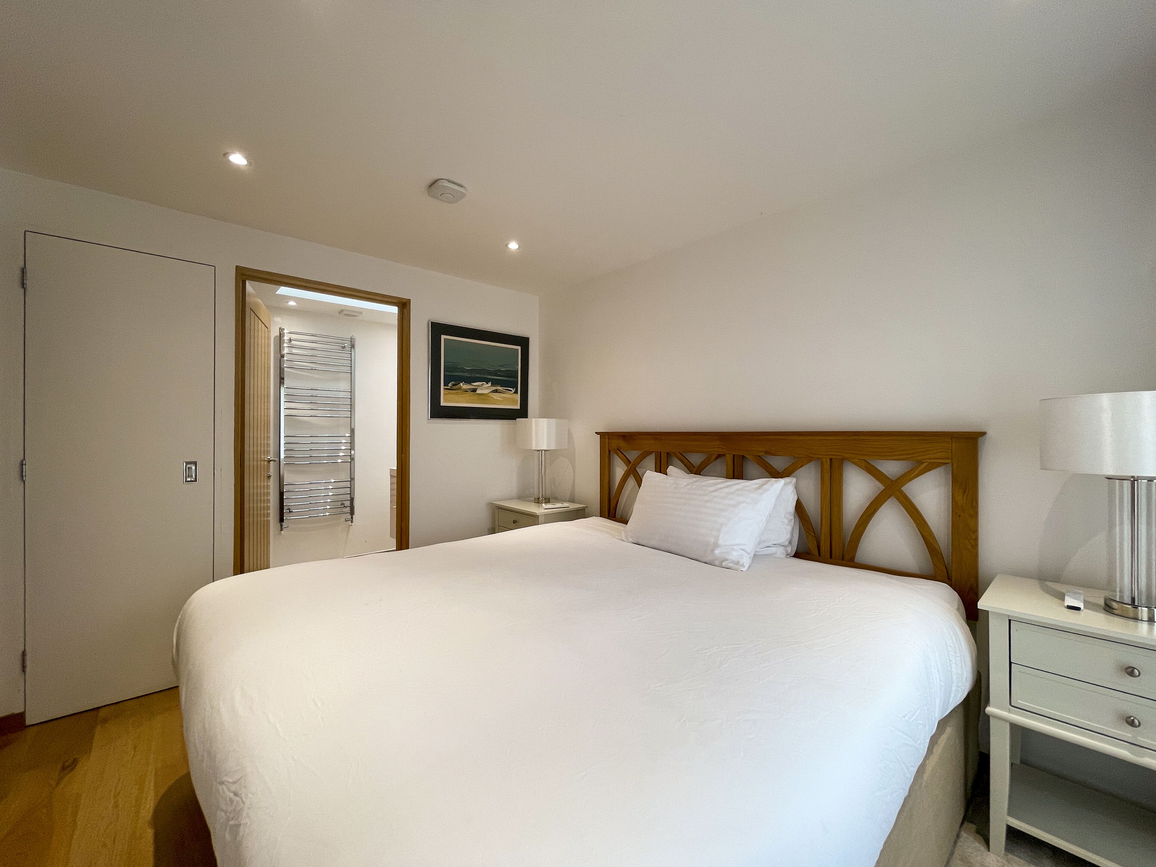 A neatly arranged bedroom with a large bed, a bedside table with a lamp, a small bedside table with a lamp, and a framed picture on the wall.