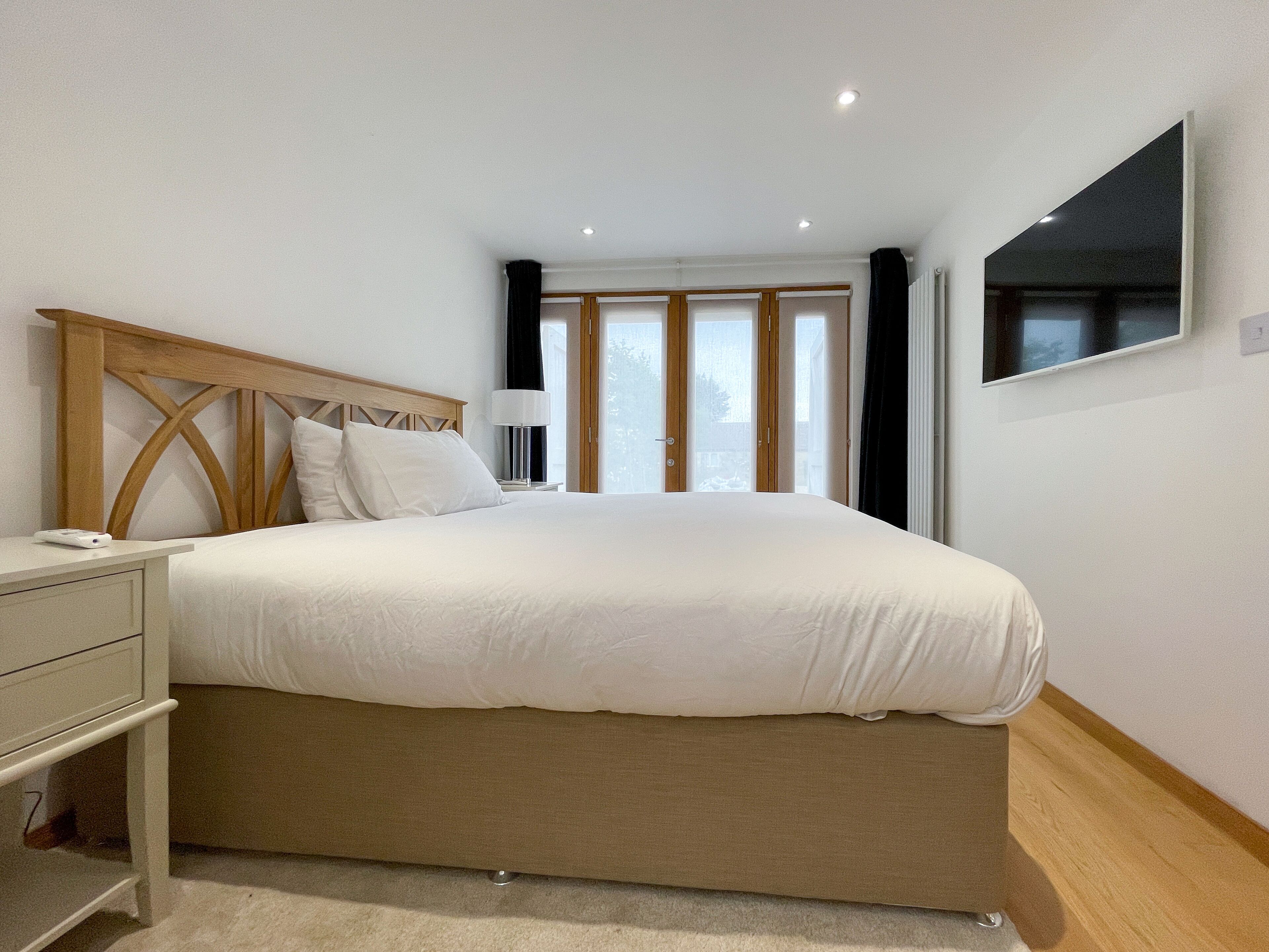 A bedroom with a large bed, a bedside table, a lamp, a TV on the wall, and a window with curtains.