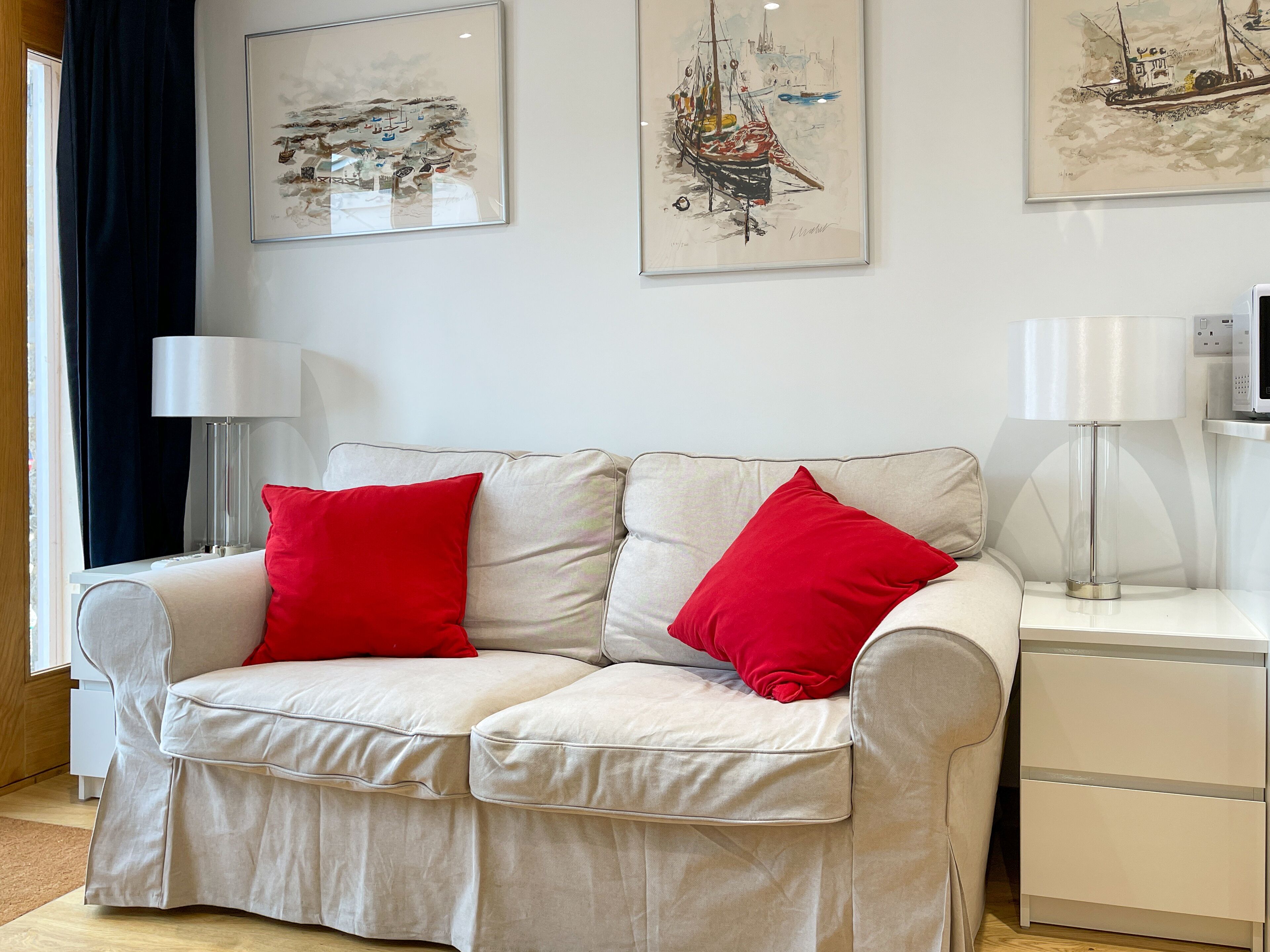 Ein modernes Wohnzimmer mit einem beigefarbenen Sofa, roten Kissen und zwei weißen Nachttischen mit Lampen.