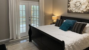 A bedroom with a bed, a nightstand with a lamp, a large floral artwork on the wall, and a view through a door with curtains. - Cozy Cottage in Rayburn Country-Pet Friendly-King Suite/Pool Table! Sleeps 10 (Brookeland)