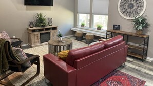 A living room with a red leather sofa, a flat-screen TV, wooden entertainment unit, and a large window with blinds. - Cozy Cottage in Rayburn Country-Pet Friendly-King Suite/Pool Table! Sleeps 10 (Brookeland)