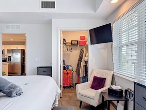 Ein Hotelzimmer mit einem Bett, einem Sessel, einem Schreibtisch, einem Fernseher und einem Kleiderschrank mit Kleidung und Taschen.