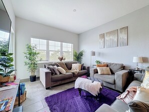 Un salon moderne avec deux canapés, une table basse et un tapis violet. On y trouve également des plantes en pot et des œuvres d’art accrochées aux murs.