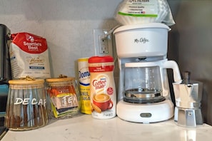 Eine Küchenarbeitsfläche mit Kaffeemaschine, Kaffeepackung, Zuckerdose, Sahnedose und einer Dose mit der Aufschrift 'DE CAF'.