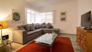 Una sala de estar con un sofá modular, una mesa de centro de vidrio, un televisor sobre un mueble de madera y obras de arte en las paredes.