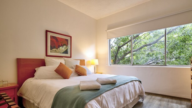 Una cama bien hecha con almohadas y una manta, una mesita de noche con una lámpara, un cuadro en la pared y una ventana con vistas a los árboles.
