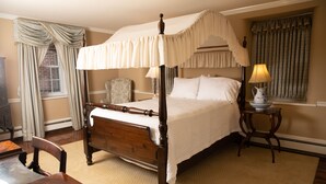 Uma cama com dossel e cortina, um criado-mudo de madeira com abajur, uma cadeira e uma cômoda.