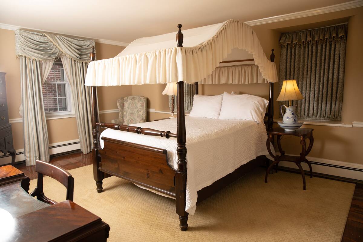 A four-poster bed with a canopy, a wooden nightstand with a lamp, a chair, and a dresser.