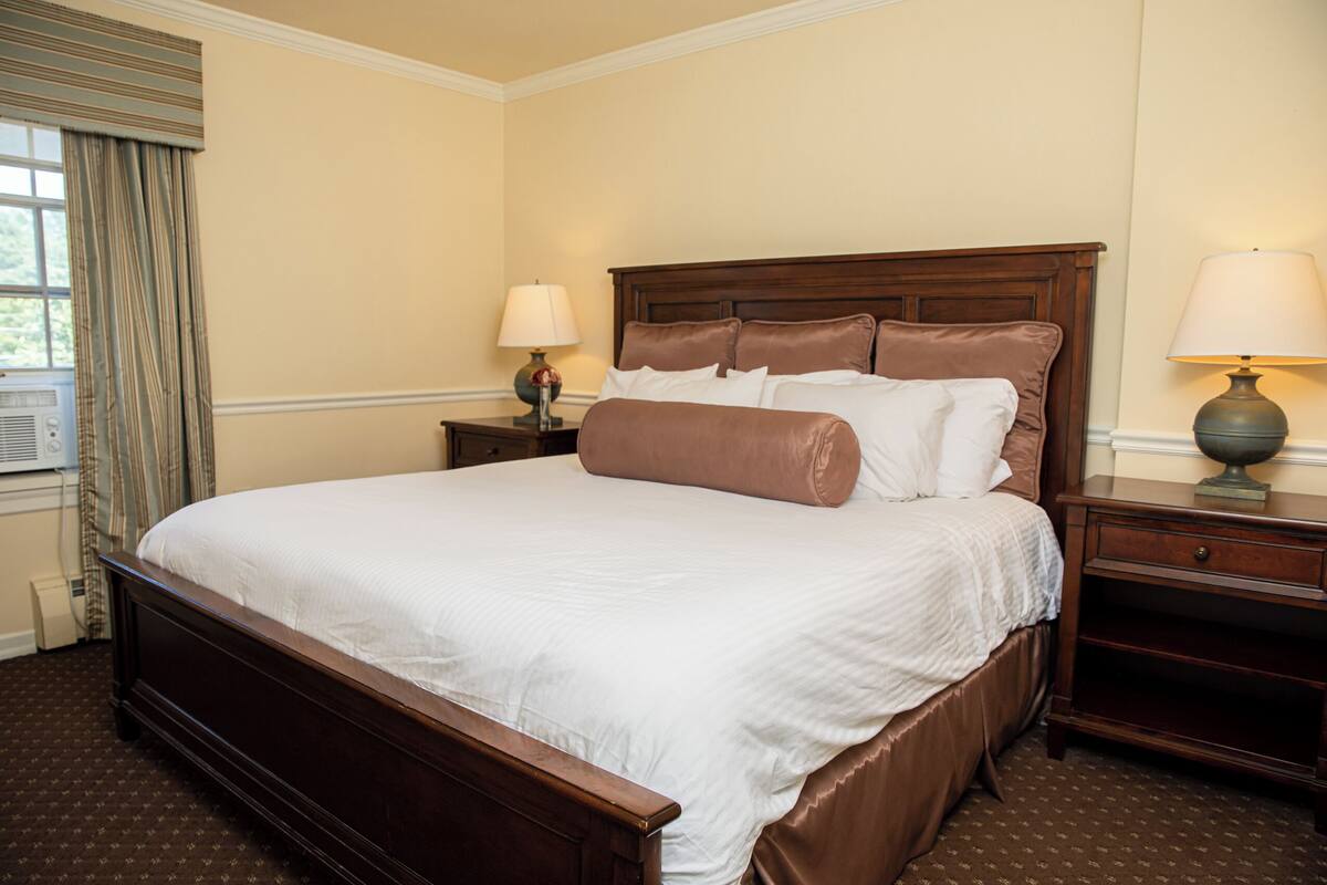 A neatly made bed with a wooden headboard, a bedside table with a lamp, and a window with curtains.