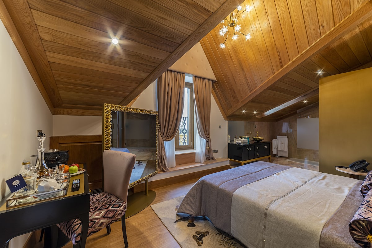 Un dormitorio con techo de madera, una cama, un escritorio con silla, una ventana con cortinas y una pequeña mesa con objetos.