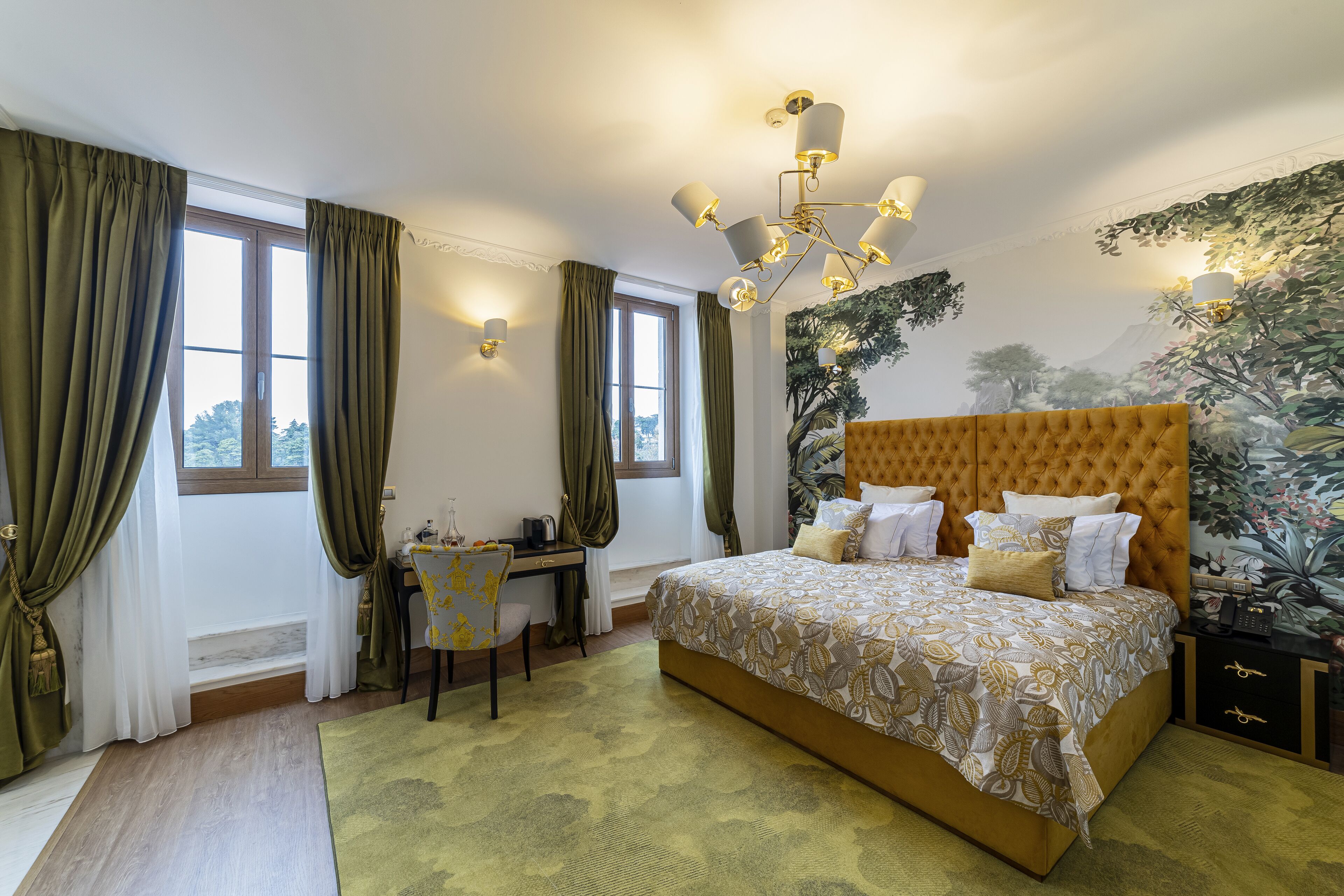 A bedroom with a large bed, a desk, and a chair. There are curtains on the windows, and a chandelier hangs from the ceiling.