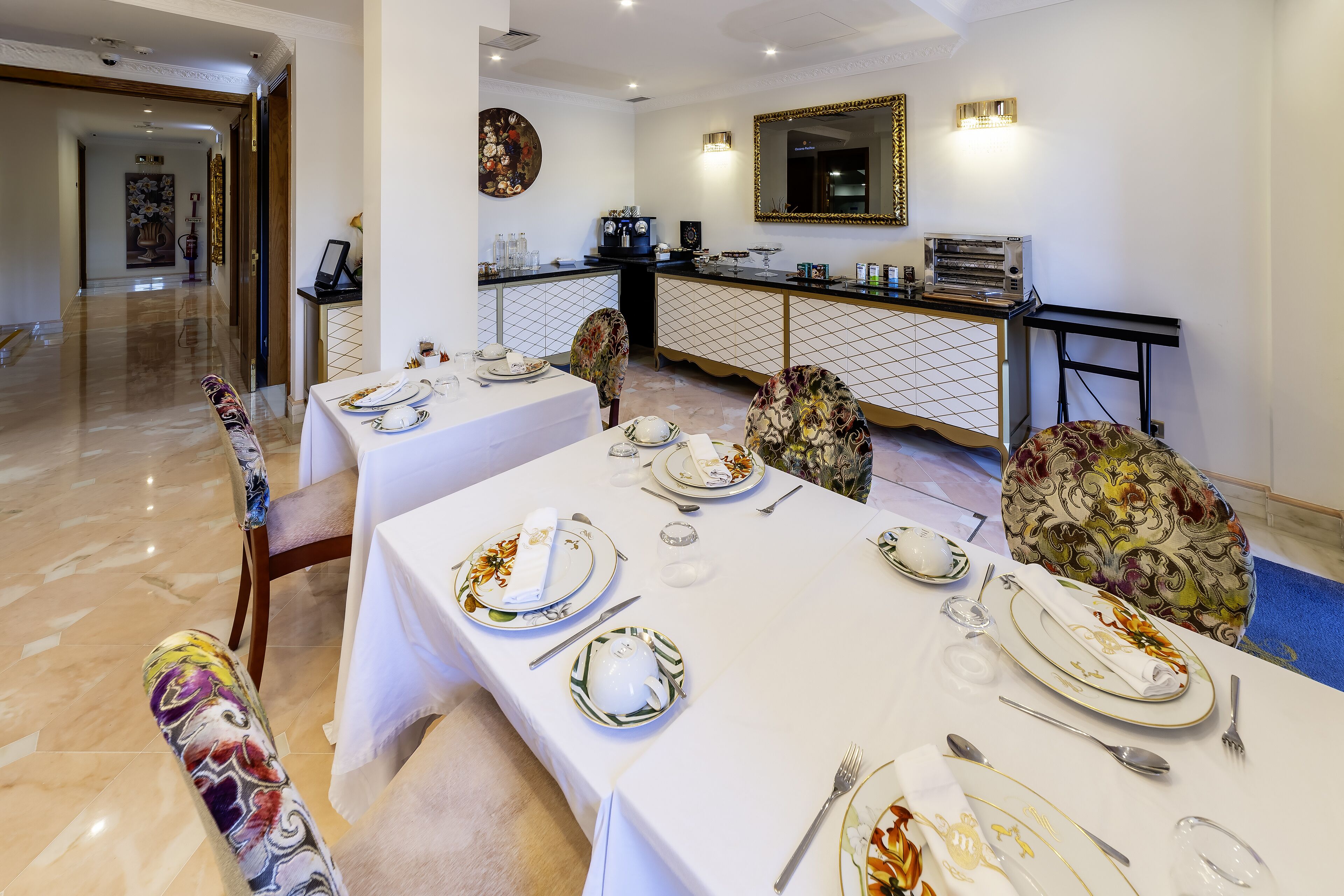A hotel dining area with tables set for service, a kitchen in the background, and a mirror on the wall.