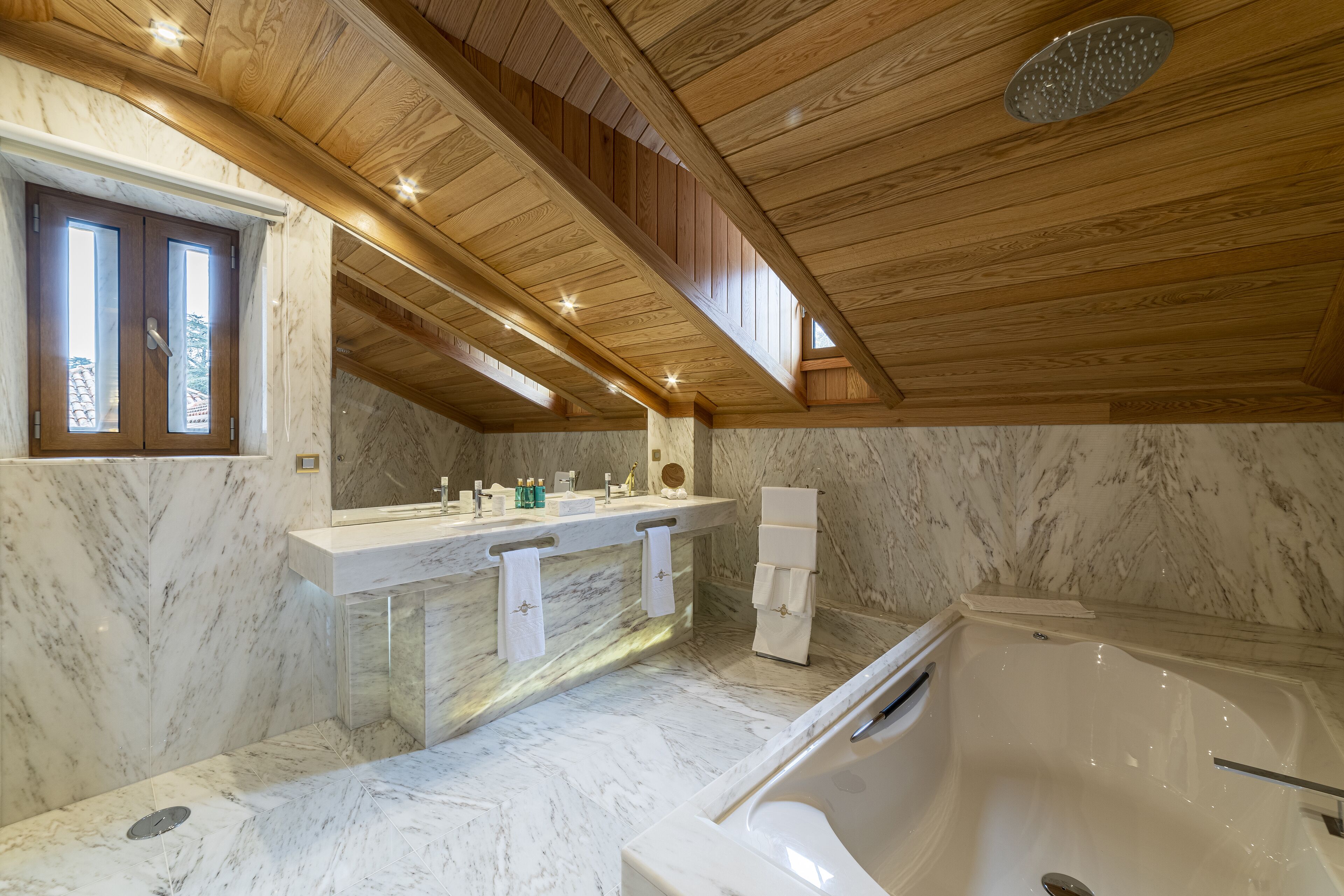 Un baño moderno con suelo de mármol, vigas de madera en el techo, una bañera grande y un tocador con espejo y toallero.