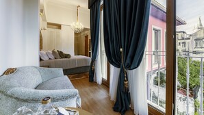 Een slaapkamer met een bed, een sofa, een kroonluchter en uitzicht op een roze gebouw door een raam met donkere gordijnen.