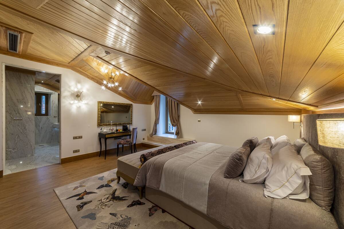 Un dormitorio con techo de madera, cama con muchos almohadones, escritorio con silla y un espejo grande.