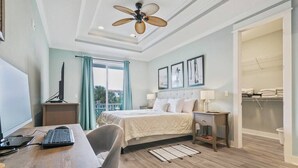 Habitación de hotel con cama, escritorio, silla y ventilador de techo. Se ve una ventana con cortinas y vistas al exterior.