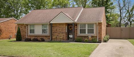 A single-story brick house with a front porch, a small lawn, and a wooden fence in the background.