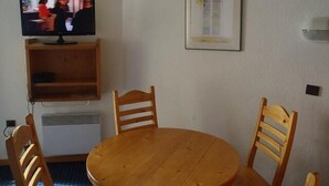 Een eetgedeelte met een ronde houten tafel, twee houten stoelen, een televisie op een houten plank, een lijst met een foto aan de muur, en een klein kastje onder de televisie.