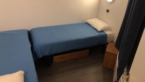 Een kleine, eenvoudige kamer met twee bedden, een houten lade onder een bed, en een klein houten kistje op de vloer.