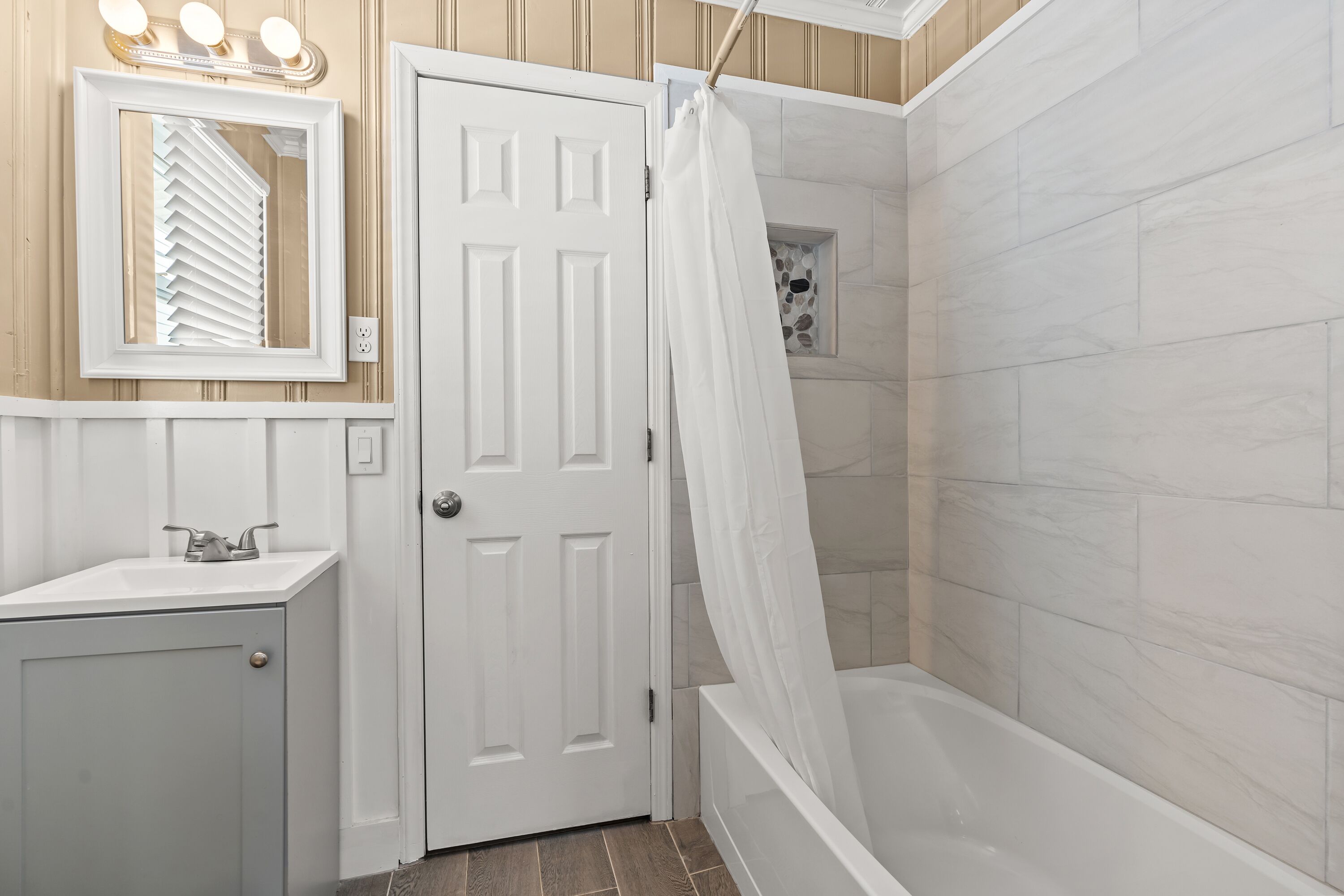 Un baño con una bañera blanca, cortina de ducha, tocador con espejo y una puerta.