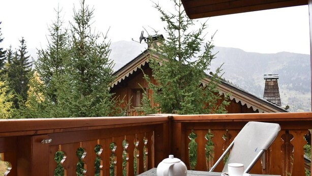 Een houten terras met een tafel en stoelen, met uitzicht op een bergachtig landschap met bomen en een chalet.