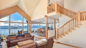 Amplia sala de estar con un gran ventanal con vistas a un lago y montañas, una escalera de madera y una zona de estar acogedora.