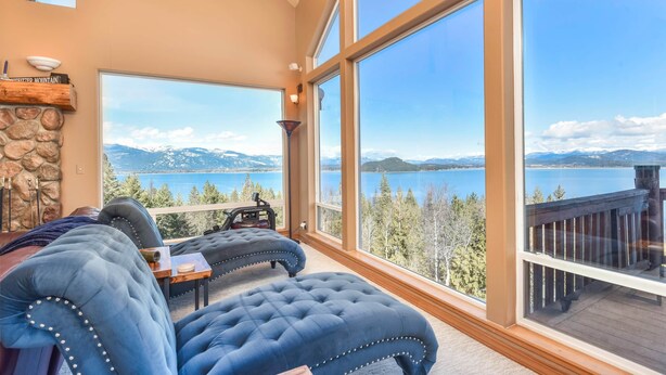 Amplia habitación con ventanas grandes que ofrecen vistas a un lago y montañas. Cuenta con una chimenea de piedra y una zona de estar cómoda con una mesa.