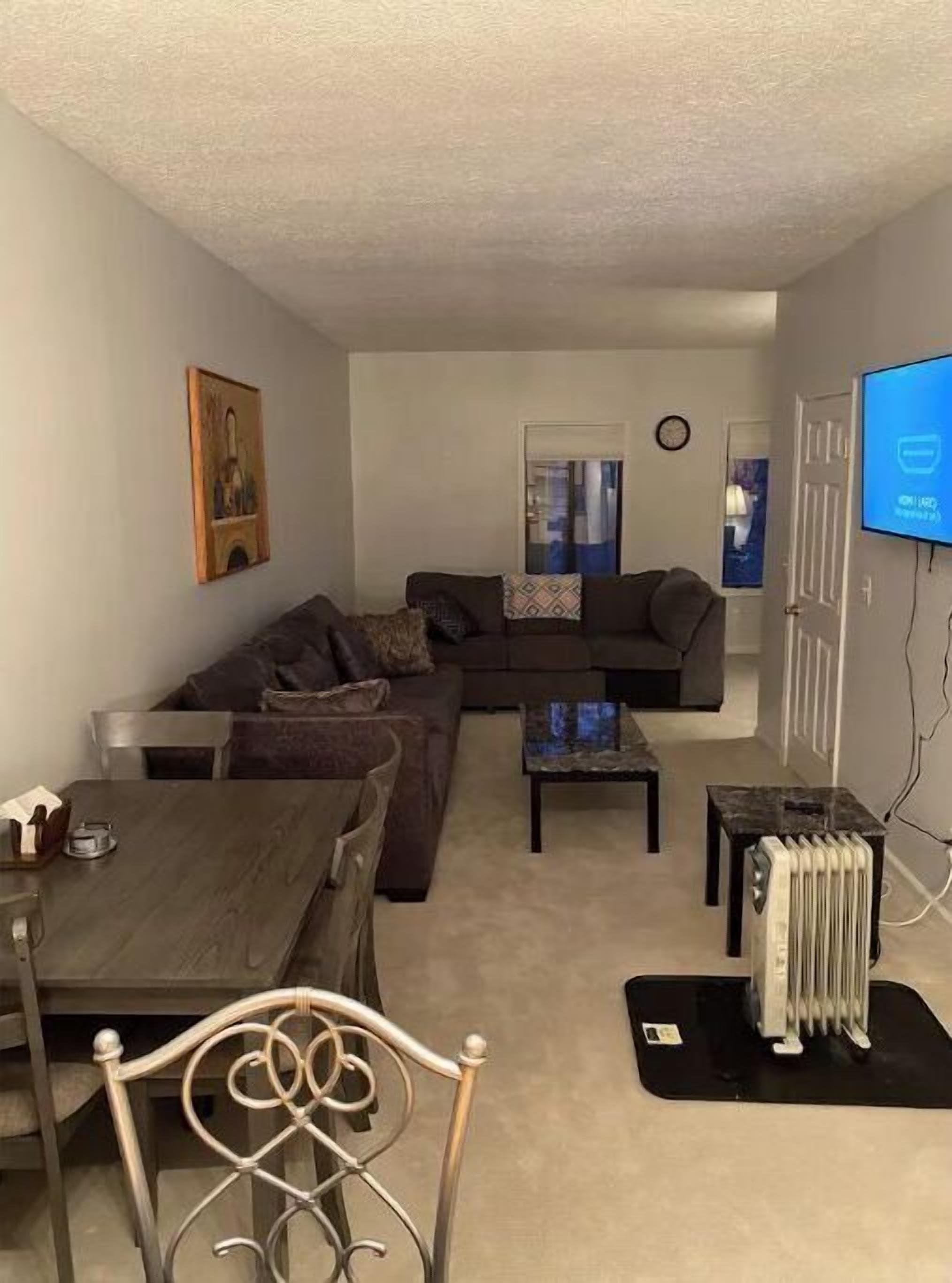 A living room with a dark brown sofa, a coffee table, and a dining table with chairs. There is a radiator on the floor and a television mounted on the wall.
