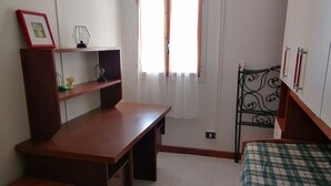 Una habitació petita i ben il·luminada amb un escriptori de fusta, un llit amb un mantell de quadres, una finestra amb cortines lleugeres i un frontal metàl·lic.