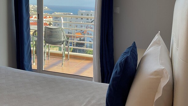 Ett hotellrum med en säng, två kuddar och utsikt över havet och byggnaderna genom ett stort fönster med gardiner.
